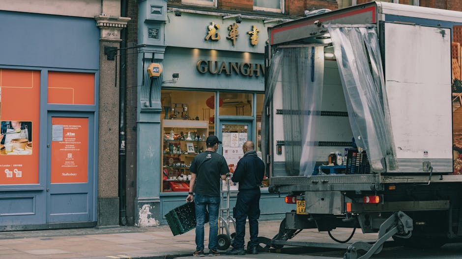 Two movers from Movers Hackney are engaged in a home relocation process outside the storefront of a shop named Guangz, which features traditional Chinese signage and decor. The movers are standing on a pavement in front of the building, with one of them holding a black plastic crate and the other managing a hand truck, both dressed in dark work clothing. A large, open-sided moving van with a transparent plastic curtain on its side is parked nearby, positioned to facilitate furniture transport and packing for the move. Inside the van, visible packing materials such as blankets and boxes are prepared for loading or unloading, with the interior showing a setup for efficient loading and transit. The area is illuminated by daylight, capturing a typical transition scene in a house removal, with objects and logistics aligned for an organized and professional move, as part of a comprehensive removals service provided by Movers Hackney, aimed at ensuring smooth home relocation or furniture transport in the Hackney area.