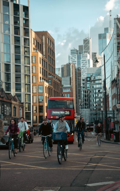 A busy urban street scene in Hackney featuring several pedestrians walking and cyclists riding their bikes, with some carrying bags or backpacks, moving along a paved area. A red double-decker bus displaying the route number 26 and destination 'Hackney Wick' is approaching in the background among modern high-rise office buildings and older brick structures, under a partly cloudy sky. The scene is well-lit with natural daylight, highlighting the mix of contemporary and historic architecture. This setting relates to house removals and home relocation services, reflecting the environment where furniture transport and packing processes might occur, as provided by Movers Hackney. The image captures the vibrant, bustling atmosphere typical of a city undergoing residential and commercial moving activities, emphasizing the logistical challenges involved in moving households within Hackney.