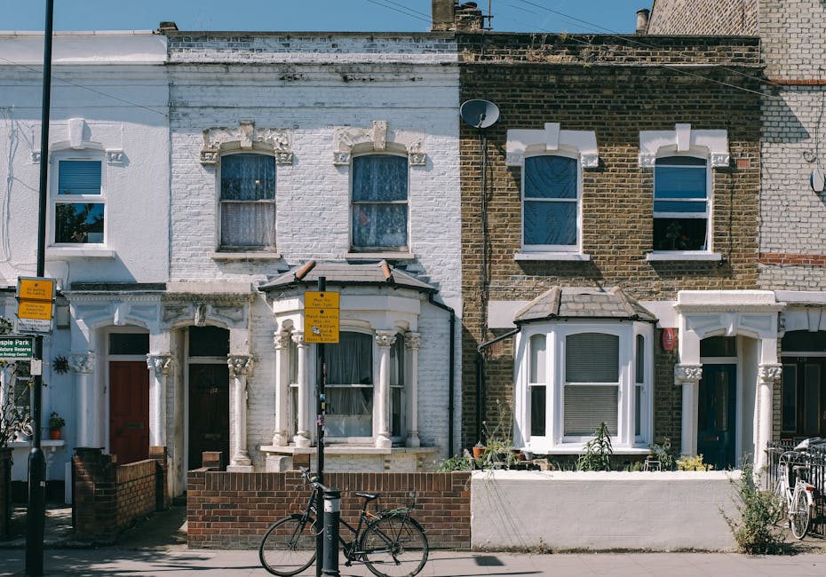 Facade of two adjacent terraced houses on Dalston Mare Street, Hackney, during a home relocation process. The house on the left has a white painted brick exterior with decorative window surrounds, a dark front door, and two upper-story windows with curtains, while the house on the right features an exposed brown brick exterior with similar windows and white decorative elements. In front of the houses, a low brick wall and a concrete planter contain a small garden with potted plants. A black bicycle is parked against a lamppost near the pavement, which has signs attached to it. The scene is captured in daylight with clear weather.