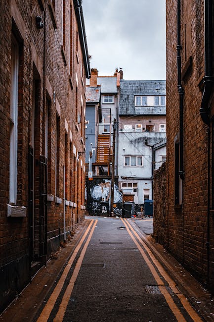 A narrow alleyway between two brick residential buildings, with a double yellow line running along the asphalt pavement. At the end of the alley, there are multi-storey buildings with visible windows, some with external staircases and fire escapes. On the pavement, there are several black trash bins and a few stacked cardboard boxes, suggesting recent or ongoing moving and packing activities. The scene is overcast, with diffused natural light highlighting the urban environment. This setting aligns with house removals and furniture transport, as it depicts the typical tight space often encountered during residential moves in Hackney, which Movers Hackney efficiently manage when providing removals services for properties like those on Dalston Mare Street.