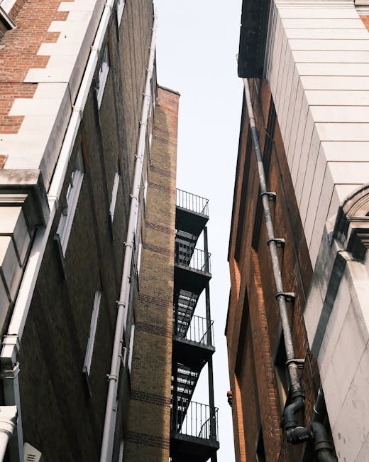 Facade of two adjacent terraced houses on Dalston Mare Street, Hackney, during a home relocation process. The house on the left has a white painted brick exterior with decorative window surrounds, a dark front door, and two upper-story windows with curtains, while the house on the right features an exposed brown brick exterior with similar windows and white decorative elements. In front of the houses, a low brick wall and a concrete planter contain a small garden with potted plants. A black bicycle is parked against a lamppost near the pavement, which has signs attached to it. The scene is captured in daylight with clear weather.