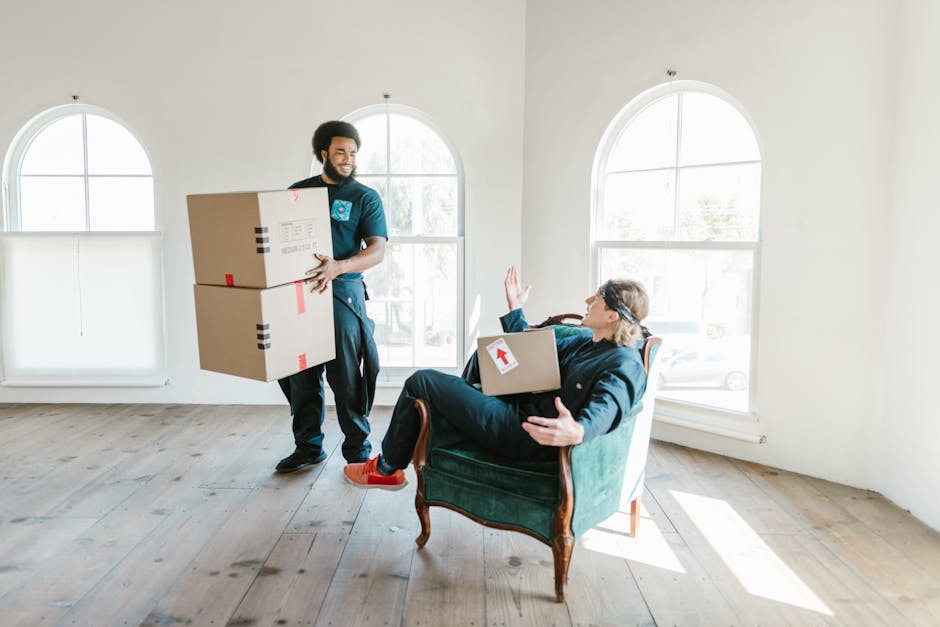 A professional mover from Movers Hackney is carrying two cardboard boxes with red and black tape markings inside a bright, spacious room with large arched windows allowing natural light to illuminate the wooden floor. The mover, dressed in dark clothing, is approaching an elderly woman seated in a vintage green upholstered armchair, who is holding a smaller cardboard box with a red arrow sticker and waving. The scene captures a home relocation or furniture transport process, with the interior environment showing minimal furnishings and an emphasis on the moving activity. The natural lighting highlights the clear, uncluttered space, reflecting the careful packing and loading stages typical of professional house removals in London. Movers Hackney's team is executing a packing and moving operation, supporting a smooth transition during house clearance or relocation efforts.