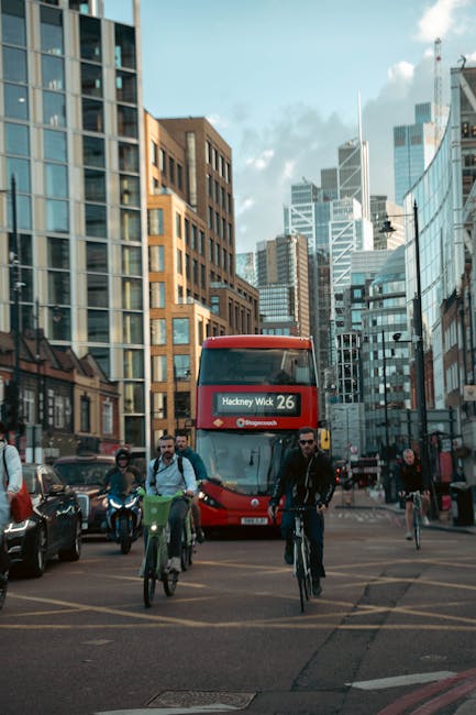A view of a busy urban street scene in London, featuring a red double-decker bus displaying 'Hackney Wick 26' on its destination board, surrounded by high-rise office and residential buildings with glass facades reflecting the sky and nearby structures. In the foreground, multiple pedestrians and cyclists navigate across a designated crossing area; some cyclists are wearing helmets and backpacks, while pedestrians include individuals dressed in casual and business attire. Several vehicles are parked along the side of the street, with a few moving in different directions. The scene is well-lit with natural daylight, highlighting the modern cityscape typical of London Fields area near Hackney, where professional removals by Movers Hackney could be relevant for home relocation or furniture transport, set against the backdrop of tall commercial buildings and a clear sky.