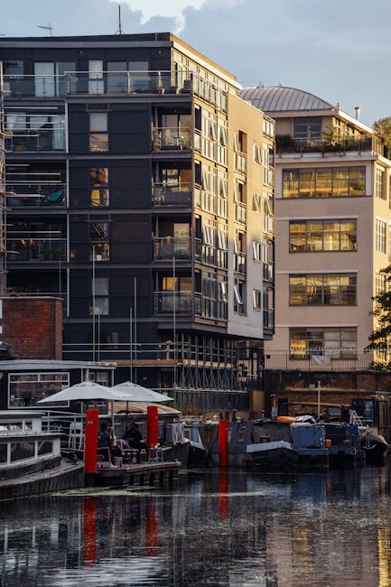 A modern multi-storey building with large glass windows and balconies, situated alongside a canal with boats moored at the water's edge. The exterior features a combination of dark and light paneling. In the foreground, there is a wooden dock with a white umbrella and seating area, and a few red bollards along the canal. Outside the building, a black van is parked, and various cardboard boxes, wrapped furniture, and moving blankets can be seen on the pavement, indicating an active house removal or moving process. Tentative signs of loading or unloading are visible as some items are stacked or being carried, reflecting a furniture transport and home relocation activity. The scene is illuminated by natural daylight, with some cloud cover, and the setting appears to be on a residential canal-side street, consistent with Hackney Wick E9 removals tips for canal side flats. Movers Hackney is likely involved in this relocation effort, supporting packing and moving services for the building’s residents.