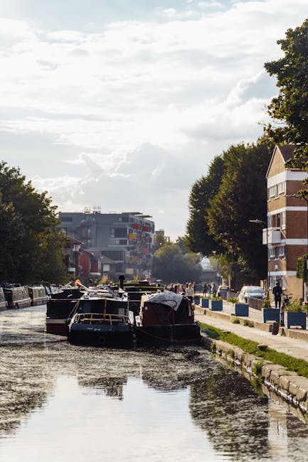 A narrow canal lined with houseboats and residential buildings visible on both sides, with a paved walkway along the water's edge. Tall trees with lush green foliage shade the path, and some pedestrians and cyclists can be seen along the sidewalk. The sky above is partly cloudy, with sunlight casting reflections on the calm water. In the background, modern apartment buildings are visible, indicating an urban residential area. The scene illustrates a typical canal-side environment in Hackney, where house removals and relocation services by Movers Hackney may be required for residents moving belongings between properties situated along the canal or nearby streets.