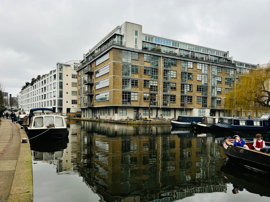 A modern multi-storey building with large glass windows and balconies, situated alongside a canal with boats moored at the water's edge. The exterior features a combination of dark and light paneling. In the foreground, there is a wooden dock with a white umbrella and seating area, and a few red bollards along the canal. Outside the building, a black van is parked, and various cardboard boxes, wrapped furniture, and moving blankets can be seen on the pavement, indicating an active house removal or moving process. Tentative signs of loading or unloading are visible as some items are stacked or being carried, reflecting a furniture transport and home relocation activity. The scene is illuminated by natural daylight, with some cloud cover, and the setting appears to be on a residential canal-side street, consistent with Hackney Wick E9 removals tips for canal side flats. Movers Hackney is likely involved in this relocation effort, supporting packing and moving services for the building’s residents.