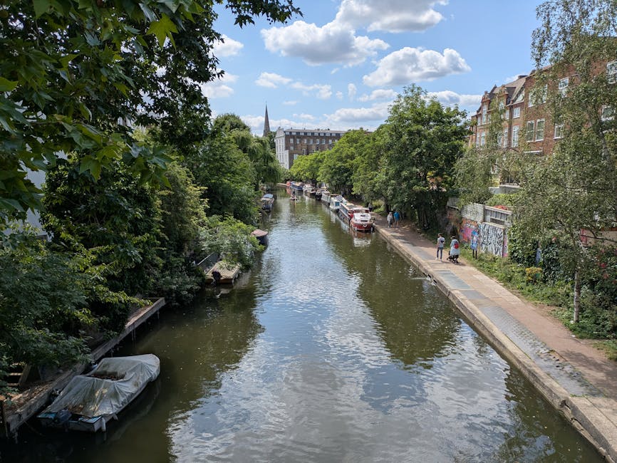 A canal scene in a residential area of Stoke Newington Church Street during daytime, with calm water reflecting a partly cloudy sky. On the left side of the canal, dense green foliage and trees extend over the water, with a small boat covered by a protective tarp moored along the bank. On the right side, a narrow concrete pavement runs parallel to the water, with a few pedestrians walking and standing nearby. Several parked cars are visible further along the street, with residential buildings featuring brick facades and large windows lining the right side of the street. In the background, a church spire rises above the trees and buildings, suggesting a historical setting. The scene captures a typical urban residential environment conducive to home relocation and furniture transport activities, with natural light illuminating the area, and the surroundings providing an inviting context for moving or packing and moving services offered by Movers Hackney.