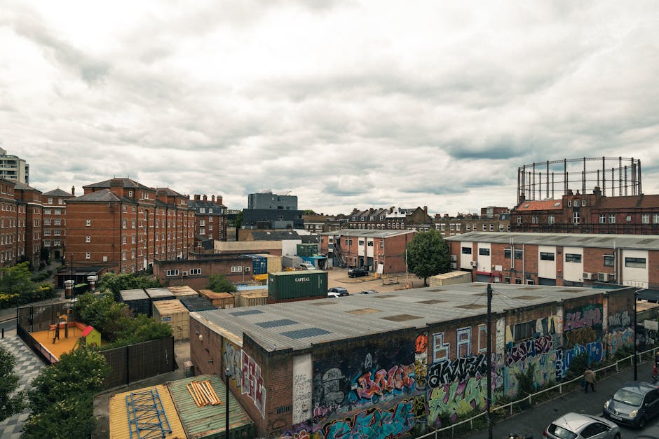 A cityscape view captured from a higher vantage point during daytime, showing a mix of residential and industrial buildings with brick and concrete facades. In the foreground, there is a building with a flat roof covered in graffiti art, adjacent to a small outdoor area with trees and a playground. Behind it, a parking lot contains several cars and a few shipping containers marked with the label 'CAPITAL.' To the right, there is a large metal structure resembling a water tower or industrial tank surrounded by scaffolding. Multiple multi-storey brick residential buildings are visible further back, with some featuring chimney stacks. The skyline is dominated by a cloudy sky with patches of grey clouds, creating diffused lighting across the scene. This urban environment reflects a typical backdrop for house removals and furniture transport, often involving careful packing and logistics, as seen in the surroundings managed by Movers Hackney for moving services on Stoke Newington Church Street.